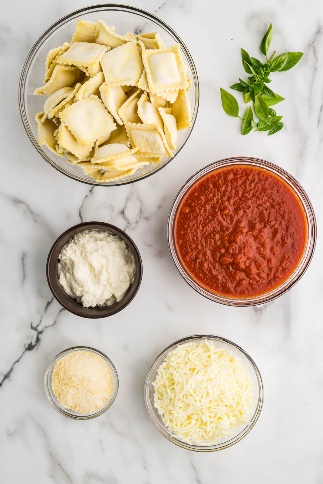 ravioli lasagna ingredients to include bowls of ravioli, spaghetti sauce, mozzarella cheese, Parmesan cheese, ricotta cheese and fresh basil