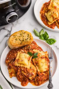 overhead view of two plates of cooked ravioli lasagna with a fork and garlic bread, with black slow cooker in the background