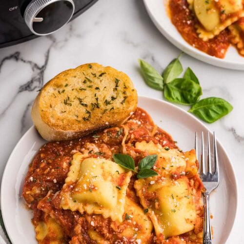 overhead view of two plates of cooked ravioli lasagna with a fork and garlic bread, with black slow cooker in the background