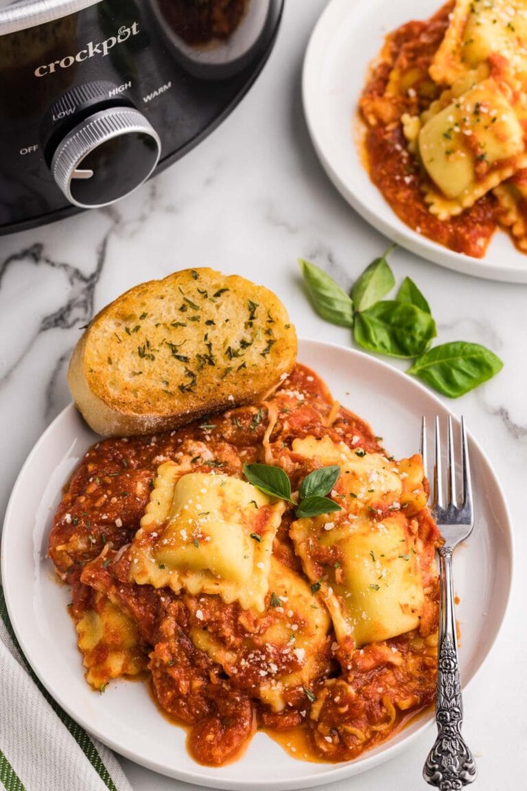 overhead view of two plates of cooked ravioli lasagna with a fork and garlic bread, with black slow cooker in the background
