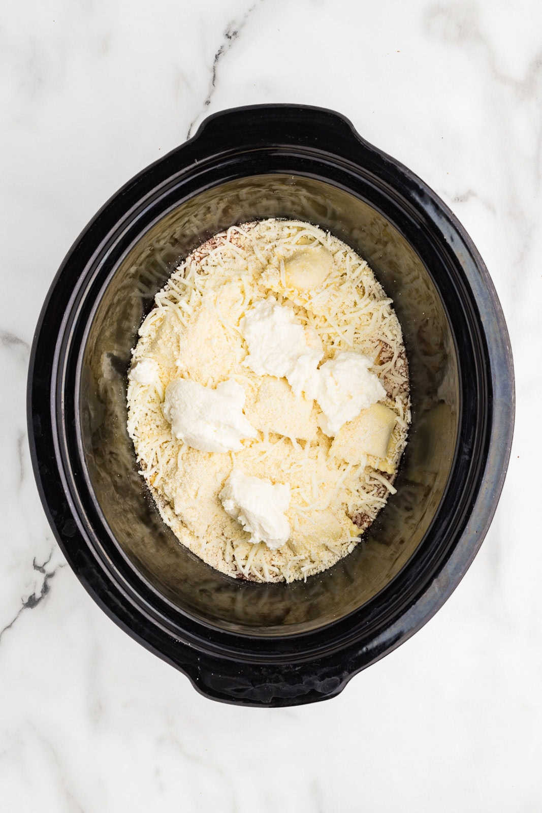 overhead view of black slow cooker with spaghetti sauce and ravioli covered by Parmesan cheese and ricotta cheese