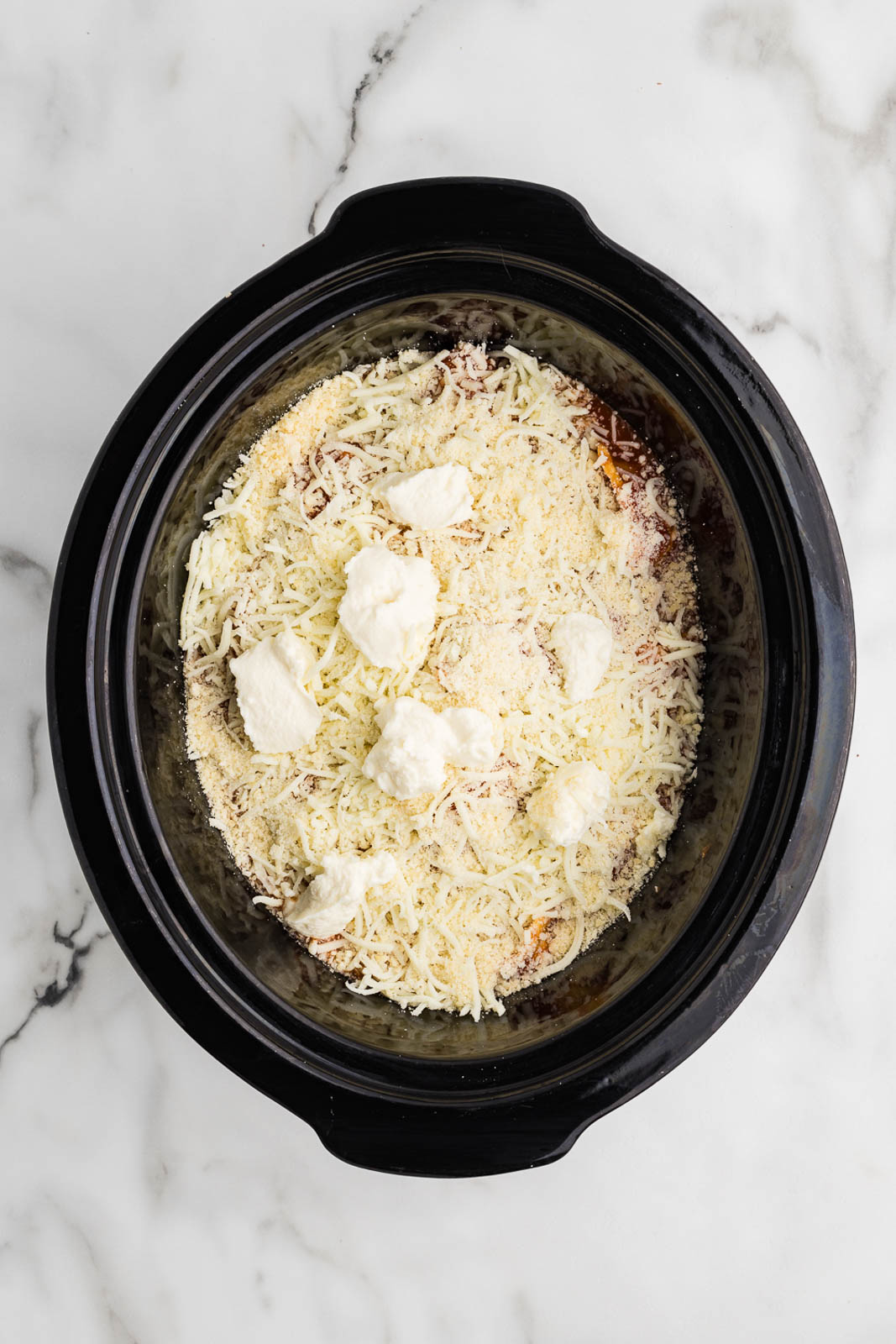 overhead view of black slow cooker with 2 layers of spaghetti sauce and ravioli covered by Parmesan cheese and ricotta cheese