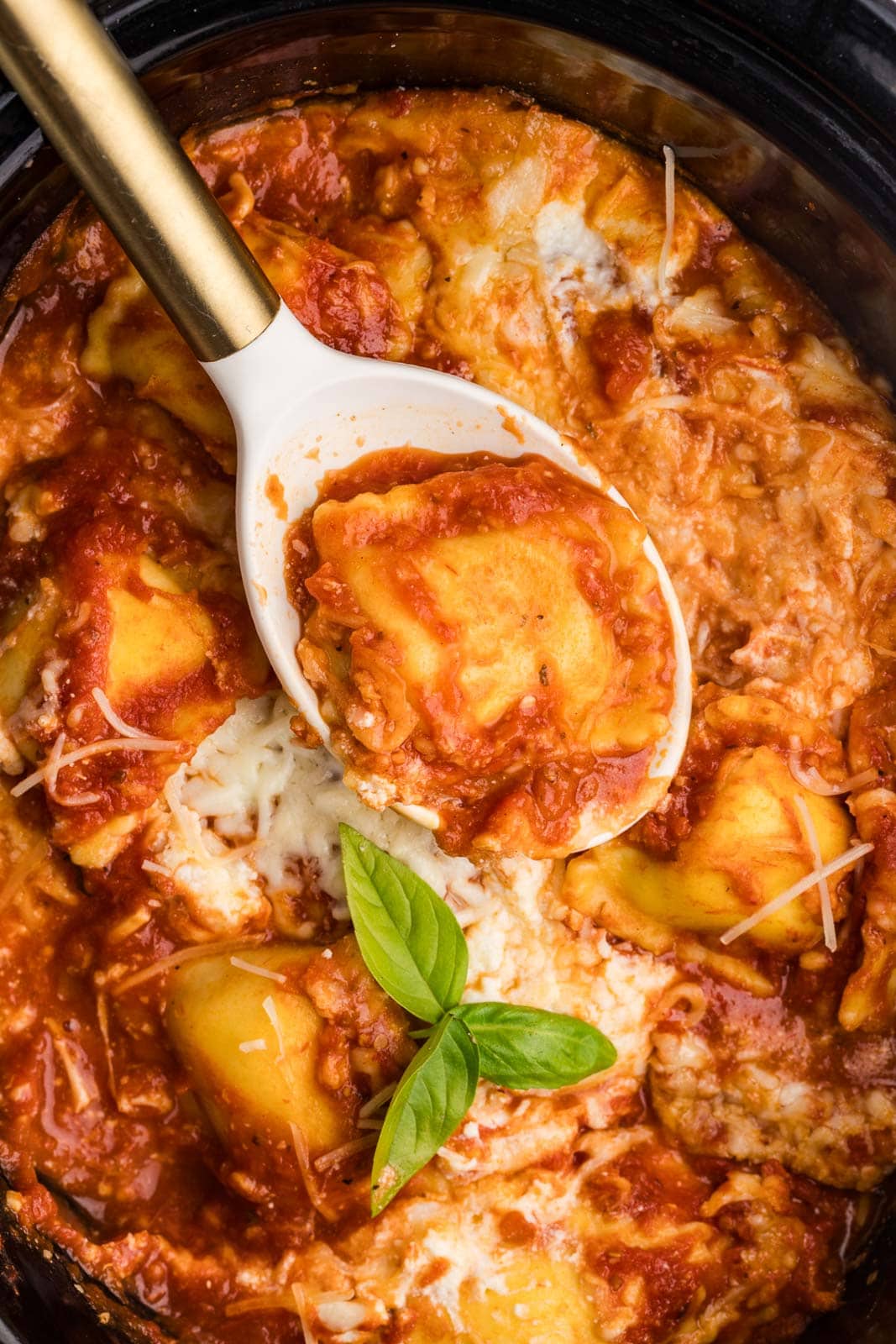 overhead view of black slow cooker with large white spoon with gold handle containing ravioli with cooked ravioli lasagna in background