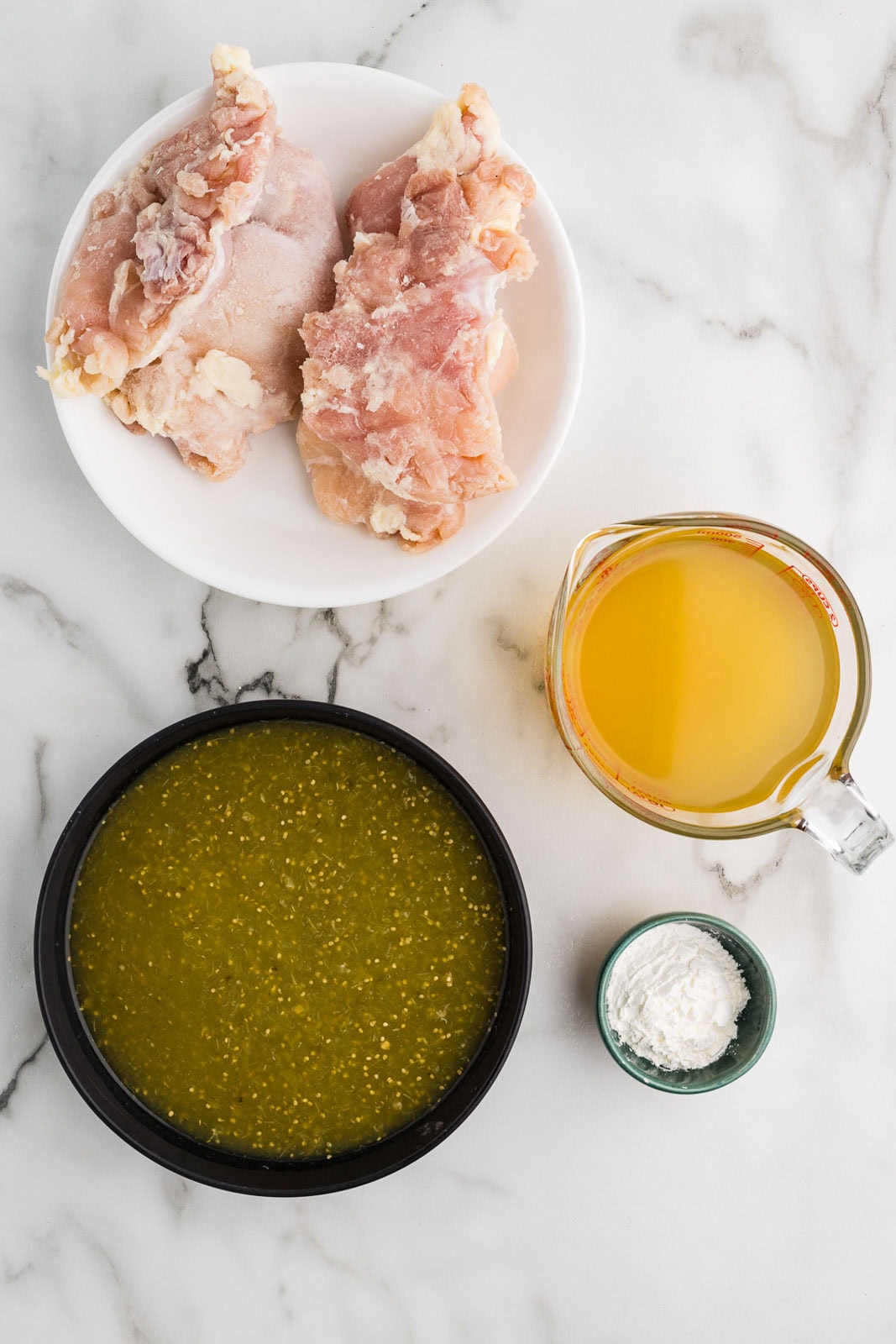 chile verde ingredients to include 2 chicken thighs on white plate, black bowl of salsa Verde, glass measuring cup of chicken broth and small bowl of cornstarch