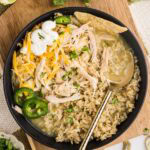 overhead view of black plate containing chile verde on rice with tortilla chips, jalapeno peppers cheese and sour cream, on a wooden cutting board with limes and cilantro in background