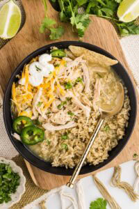 overhead view of black plate containing chile verde on rice with tortilla chips, jalapeno peppers cheese and sour cream, on a wooden cutting board with limes and cilantro in background