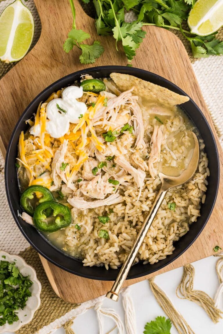 overhead view of black plate containing chile verde on rice with tortilla chips, jalapeno peppers cheese and sour cream, on a wooden cutting board with limes and cilantro in background