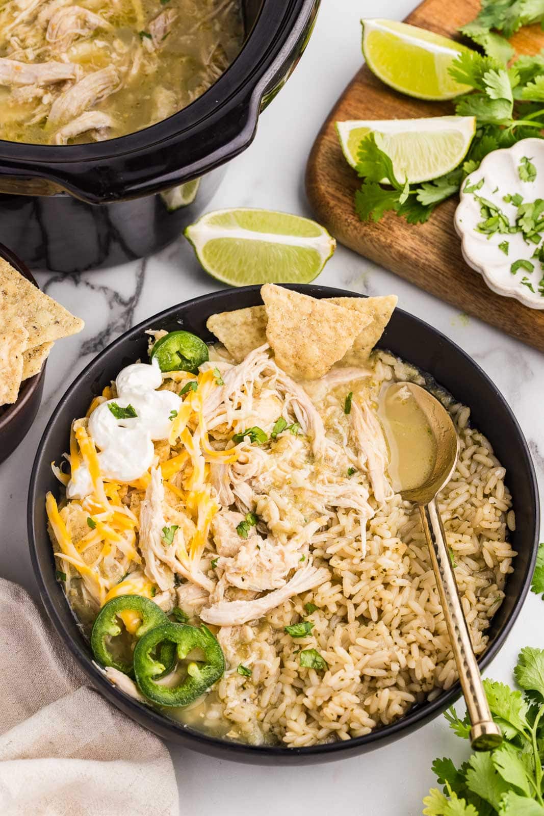 overhead view of black plate containing chile verde on rice with tortilla chips, jalapeno peppers cheese and sour cream, with limes and cilantro on a wooden cutting board with black slow cooker in background