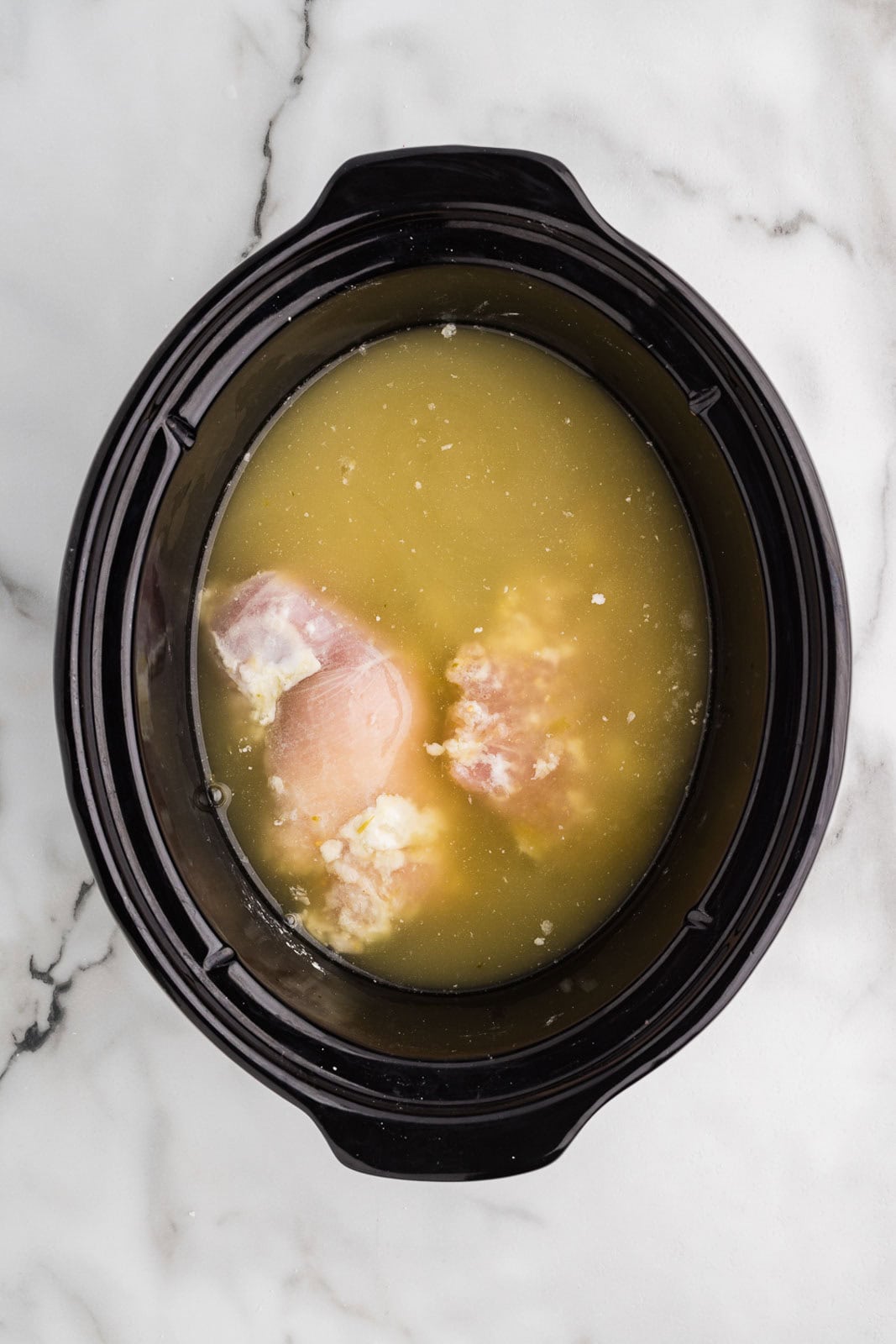 overhead view of chicken thighs in black slow cooker covered with cornstarch, salsa and chicken broth