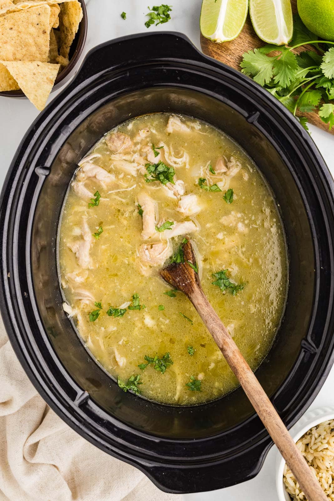 overhead view of chile verde in black slow cooker with wooden spoon inserted, surrounded by tortilla chips, limes and cilantro