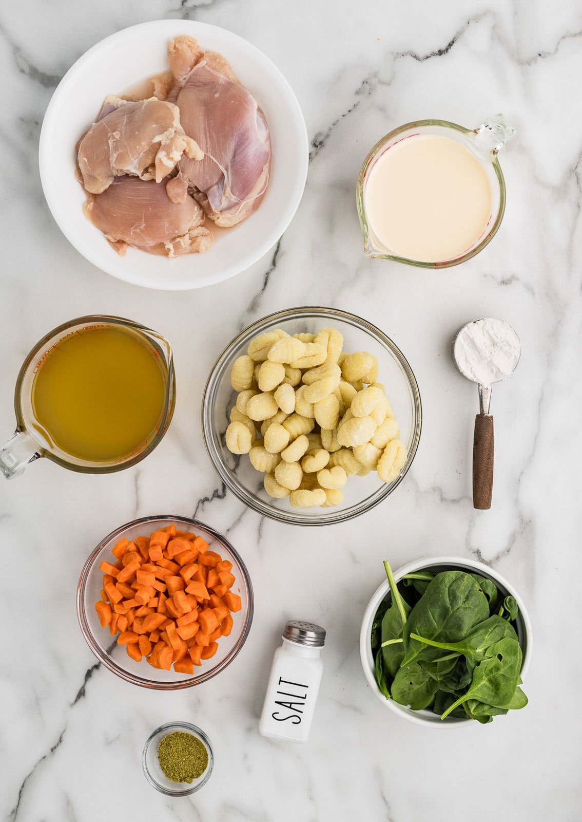 chicken gnocchi soup ingredients to include bowls of chicken thighs, gnocchi, diced carrots, spinach, herb and garlic seasoning, salt shaker, glass measuring cups of chicken broth and evaporated milk, and measuring spoon of cornstarch