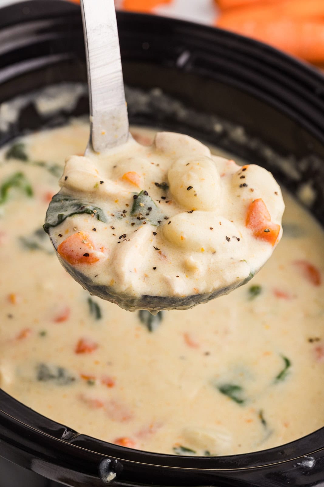 closeup of large ladle containing chicken gnocchi soup with black slow cooker of soup in the background with raw carrots on side