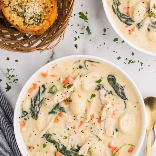 overhead view of two bowls of chicken gnocchi soup with basket of garlic bread, dishcloth and spoons on side