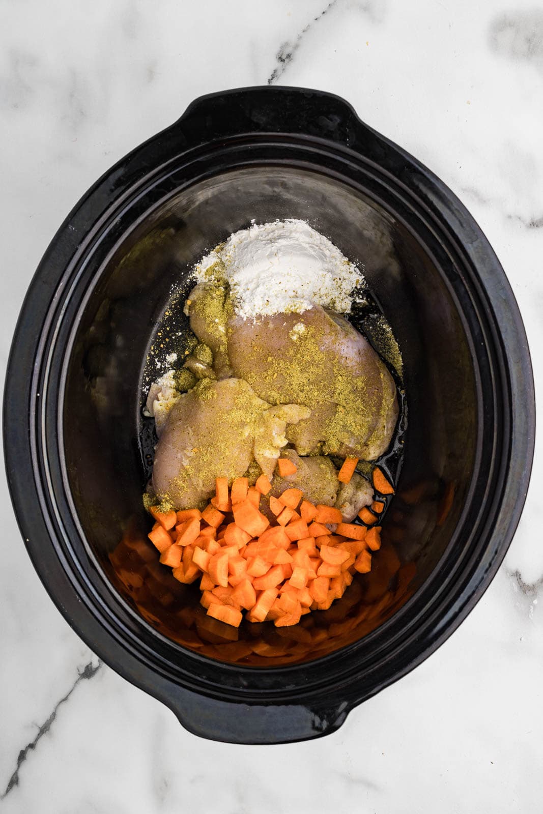 black slow cooker containing chicken thighs, carrots, cornstarch and seasoning