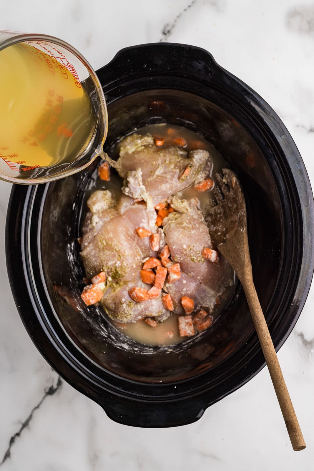 black slow cooker containing chicken thighs, carrots, cornstarch and seasoning with wooden spoon and glass measuring cup of broth being poured into slow cooker