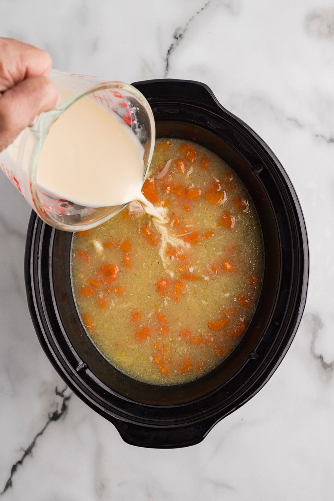 black slow cooker with cooked chicken, broth and carrots with glass measuring cup pouring evaporated milk into slow cooker