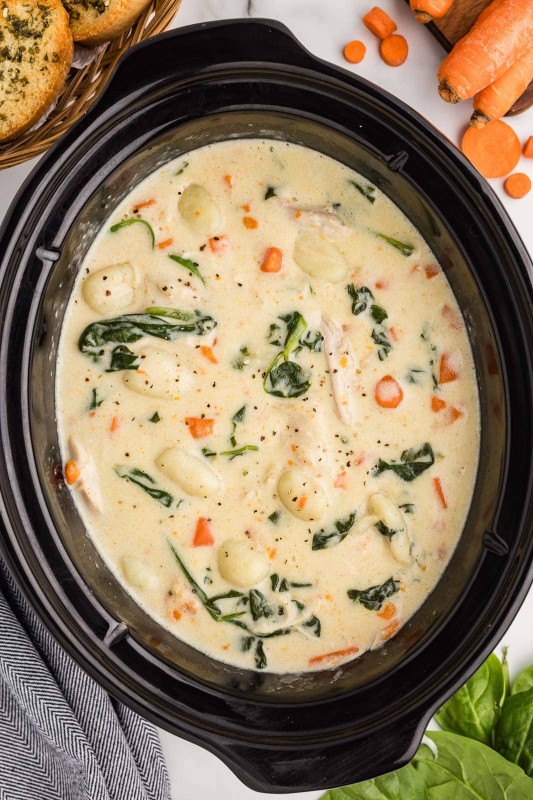 overhead view of black slow cooker with completed chicken gnocchi soup surrounded by raw carrots, basket of basket of garlic bread, raw spinach and dish towel
