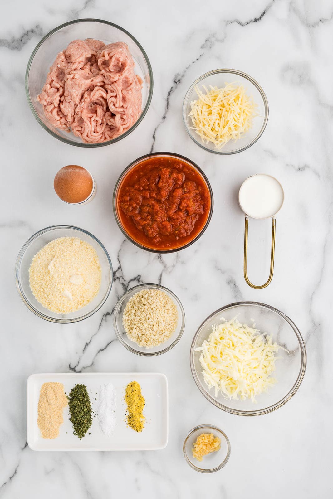chicken parmesan meatballs ingredients to include glass bowls of ground chicken, grated and shredded parmesan cheese, pasta sauce, one egg, milk, Panko breadcrumbs, mozzarella cheese, minced garlic, and plate of seasonings