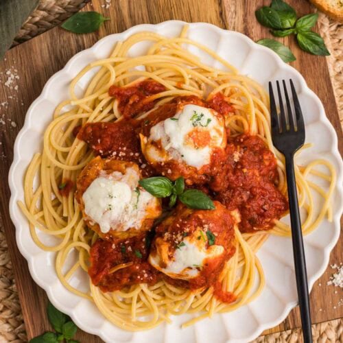 white dinner plate of meatballs over spaghetti with black fork on the side, on wooden cutting board, surrounded by placemat and portion of garlic toast