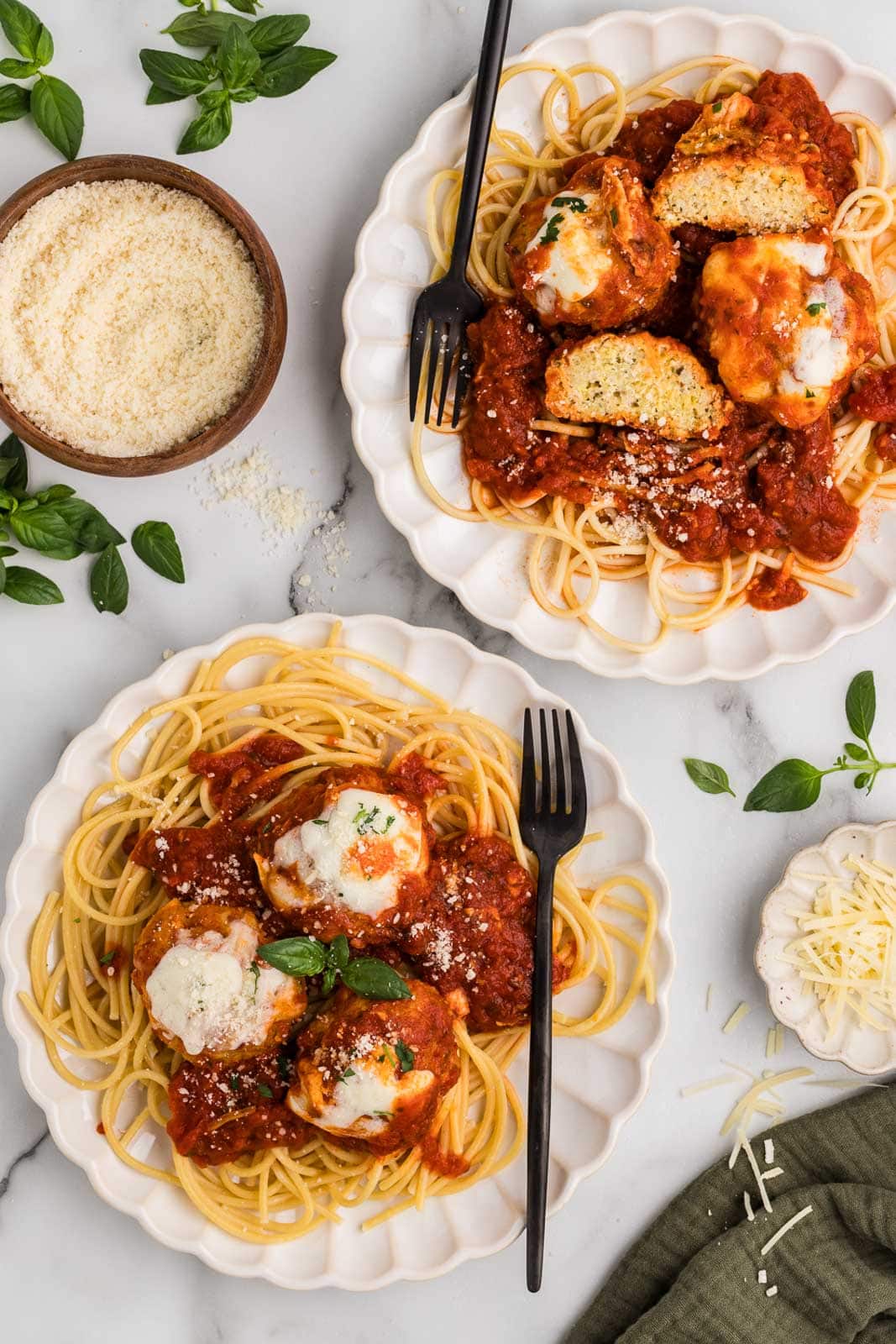 two white dinner plates of spaghetti and meatballs with black forks, with wooden bowl of grated parmesan cheese on side
