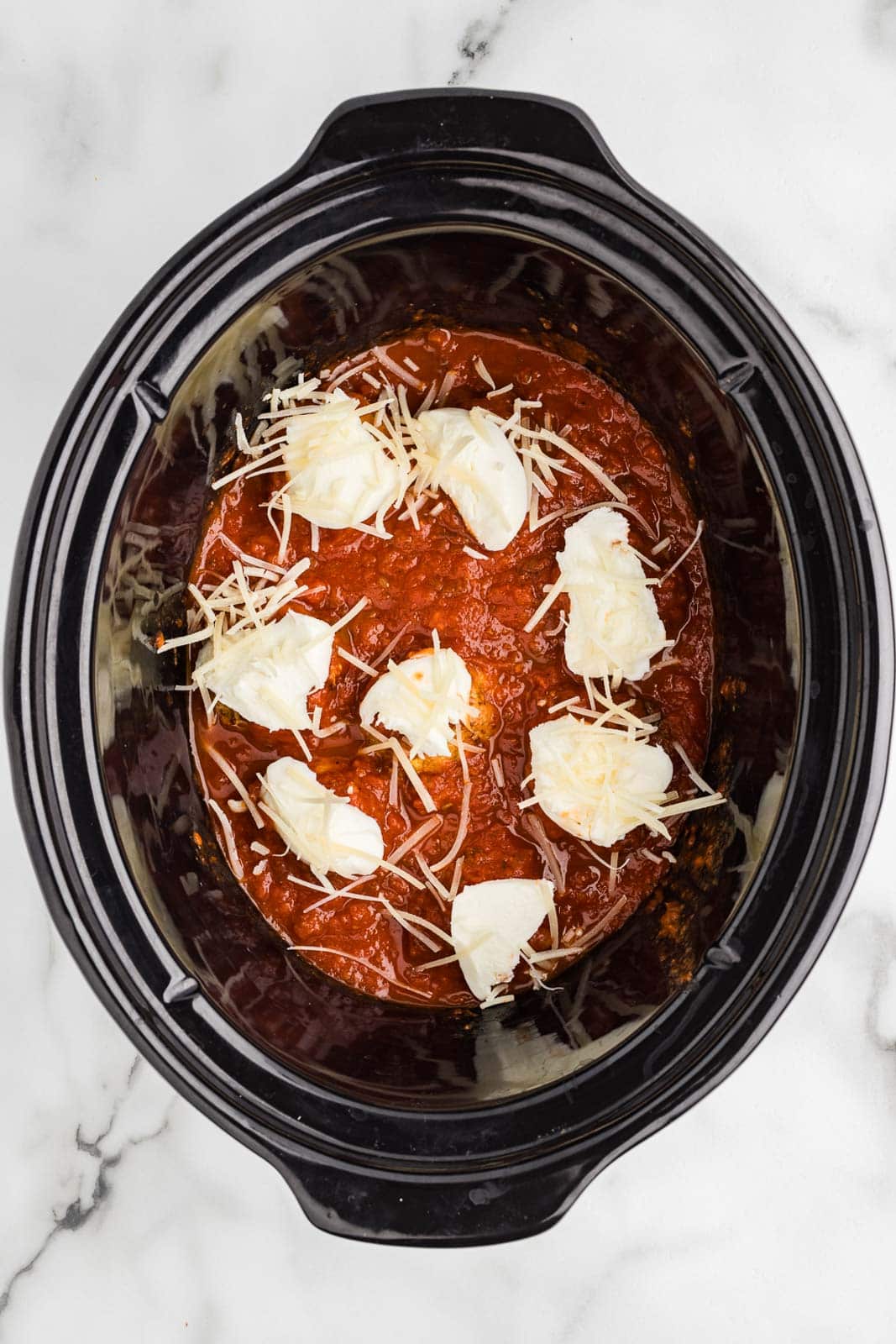 overhead view of black slow cooker with meatballs covered by pasta sauce and mozzarella cheese