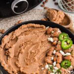 front view of black bowl of refried beans on placemat with jalapenos on top, wooden spoon and black slow cooker in background