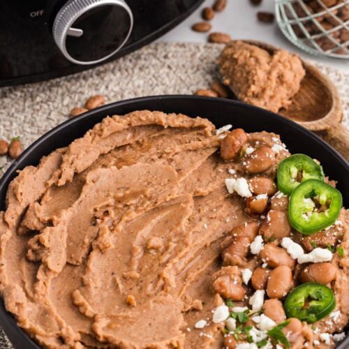 front view of black bowl of refried beans on placemat with jalapenos on top, wooden spoon and black slow cooker in background