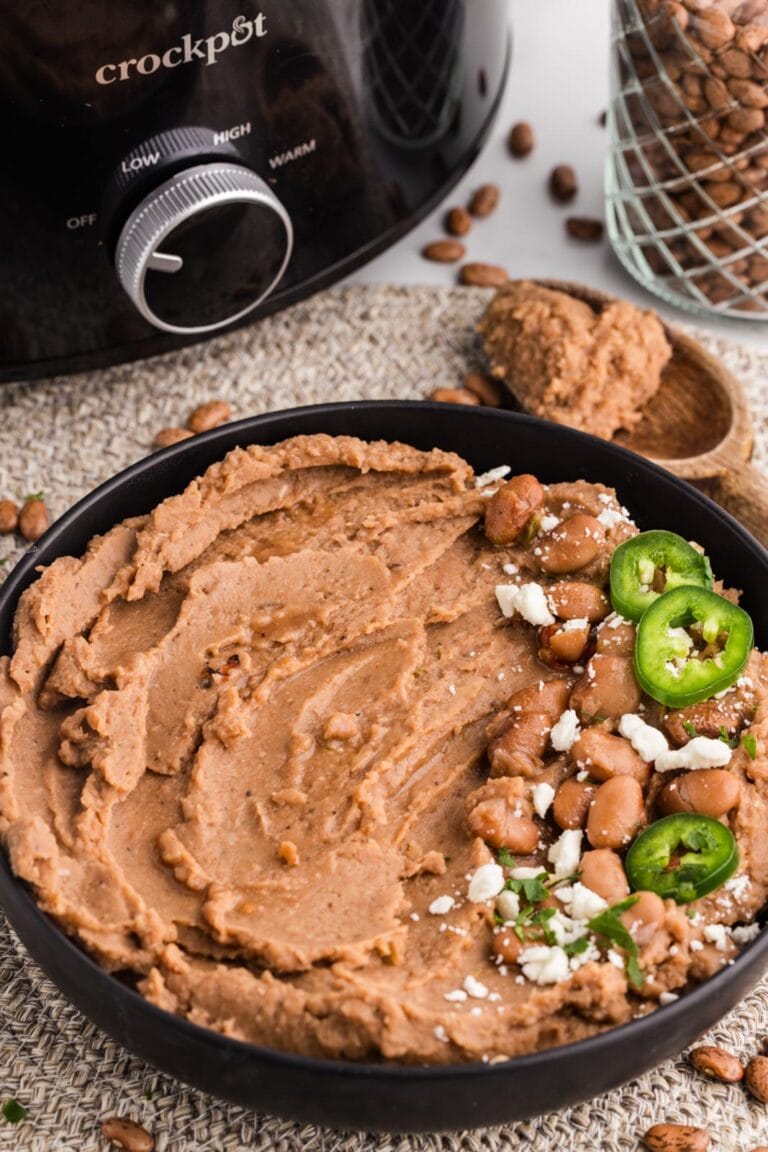front view of black bowl of refried beans on placemat with jalapenos on top, wooden spoon and black slow cooker in background
