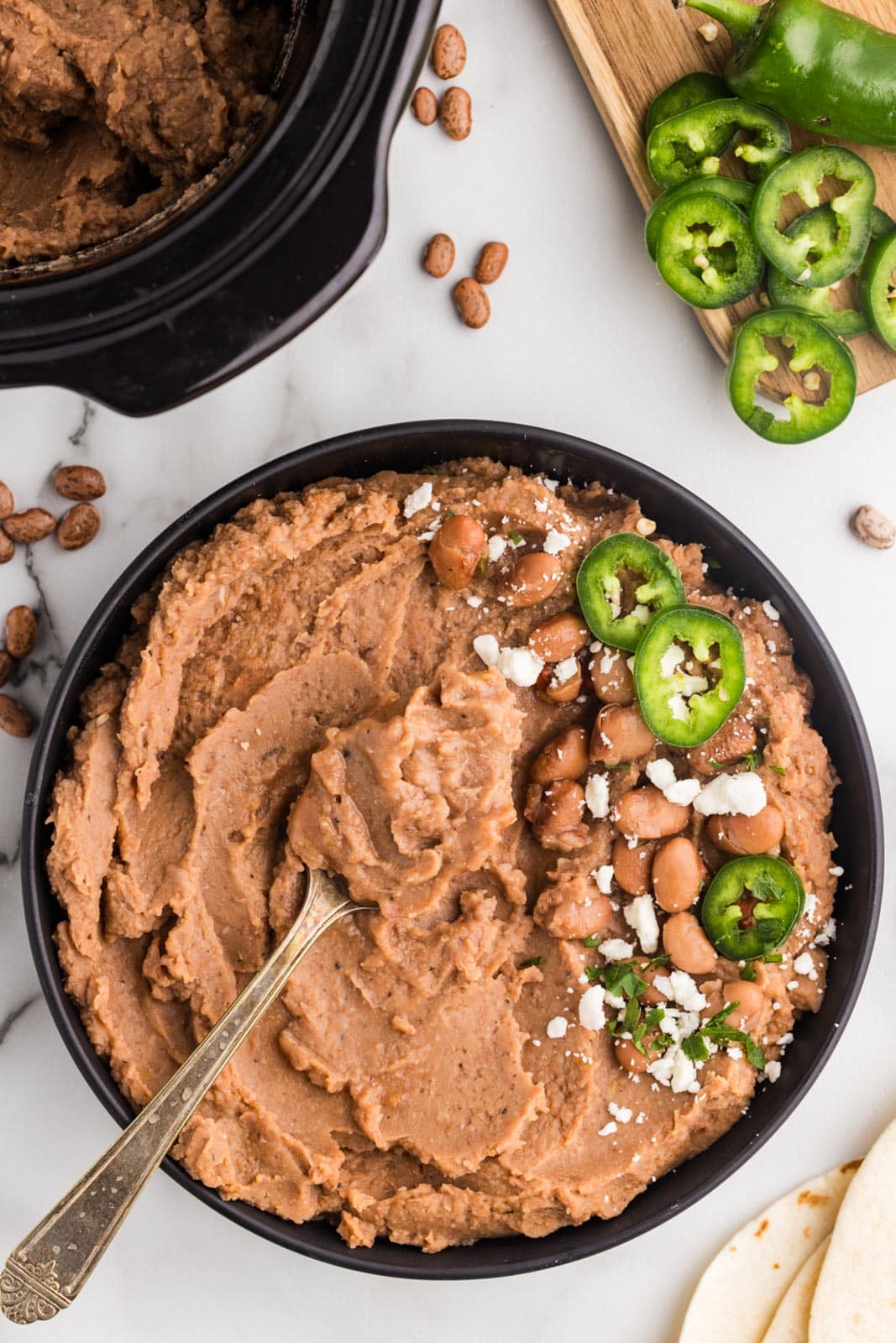 overhead view of black bowl of refried beans with jalapeno peppers on top with spoon inserted, black slow cooker and cutting board with jalapeno peppers in background