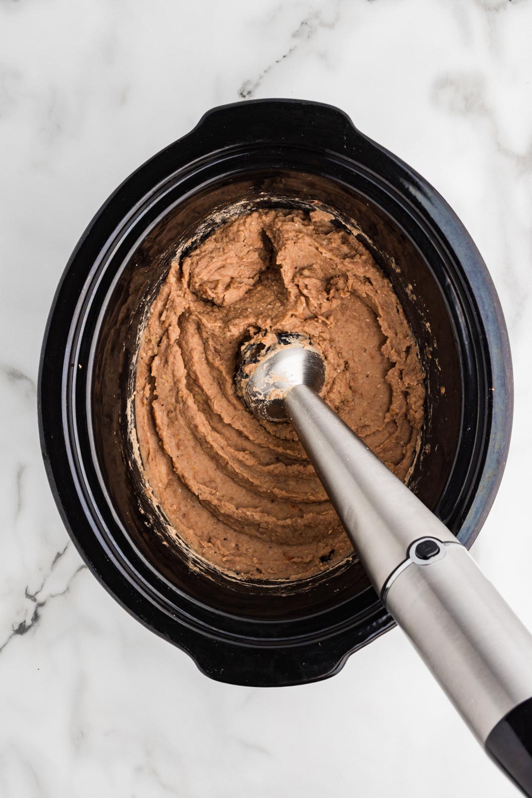 overhead view of black slow cooker with cooked beans with immersion blender