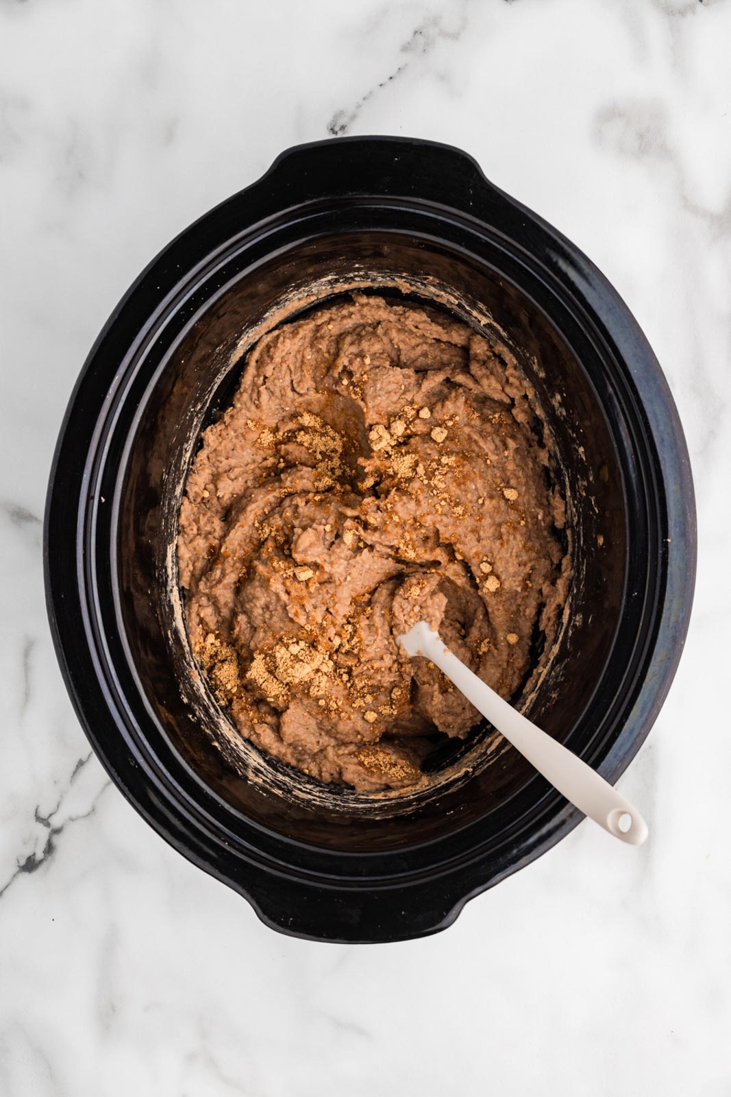 overhead view of black slow cooker of blended refried beans with white spatula