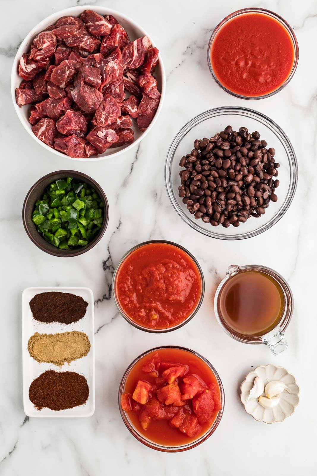 steak chili ingredients including bowls of beef stew meat, crushed tomatoes, diced tomatoes, tomato sauce, beef broth, black beans, garlic cloves, jalapeno peppers, and seasonings