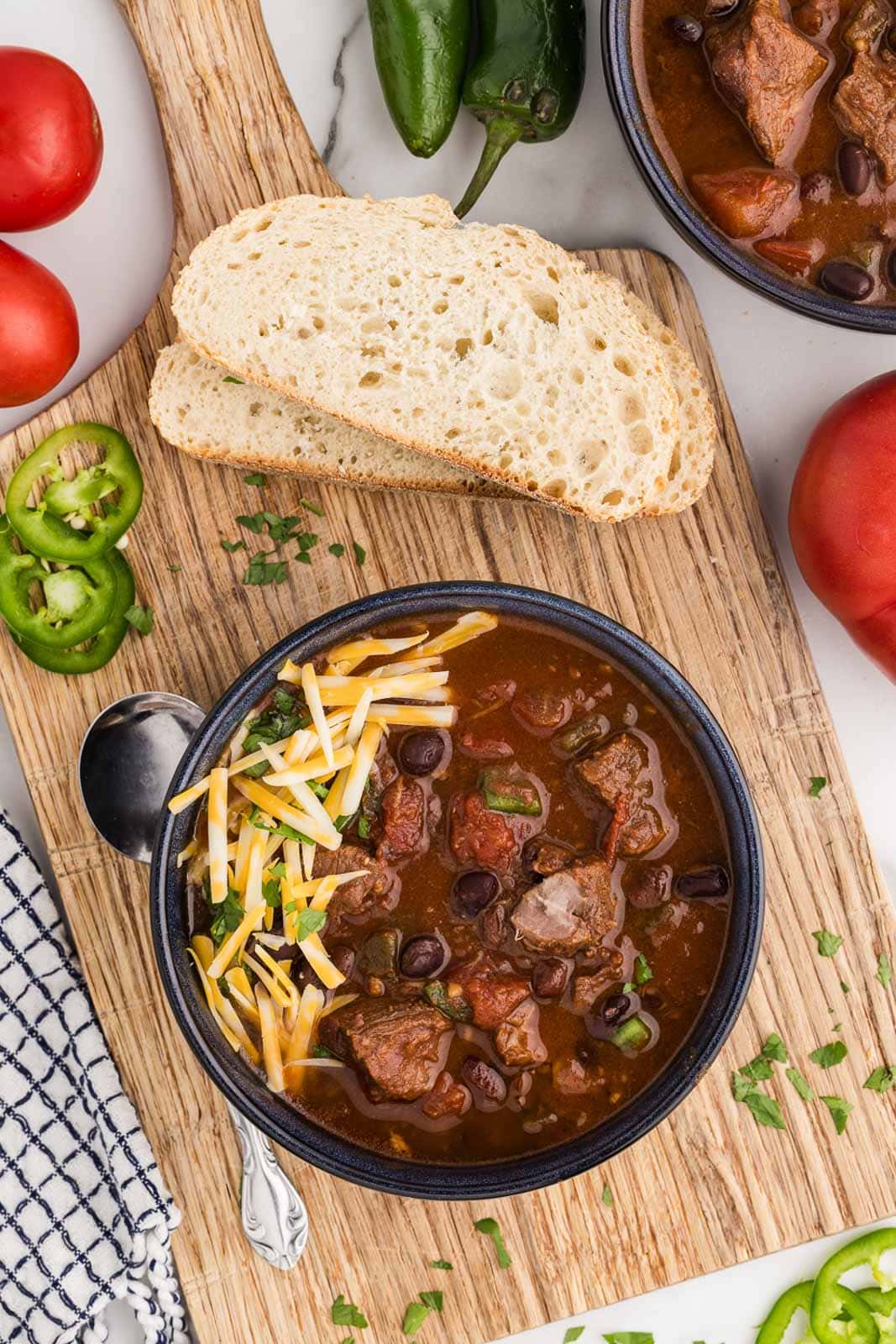 overhead view of bowl of steak chili on wooden cutting board with spoon, two pieces of bread and peppers on the side, surrounded by another bowl of chili, tomatoes, peppers dishcloth on side