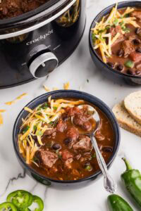 overhead view of two bowls of chili with black slow cooker in background, surrounded by sliced bread and peppers on side