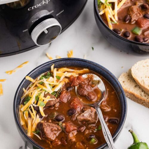 overhead view of two bowls of chili with black slow cooker in background, surrounded by sliced bread and peppers on side