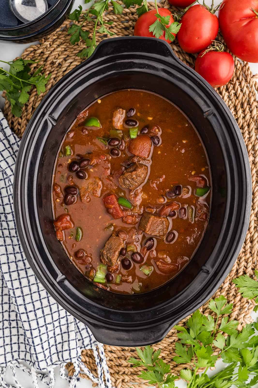 overhead view of black slow cooker with steak chili slow cooked on a placemat, with tomatoes on the side
