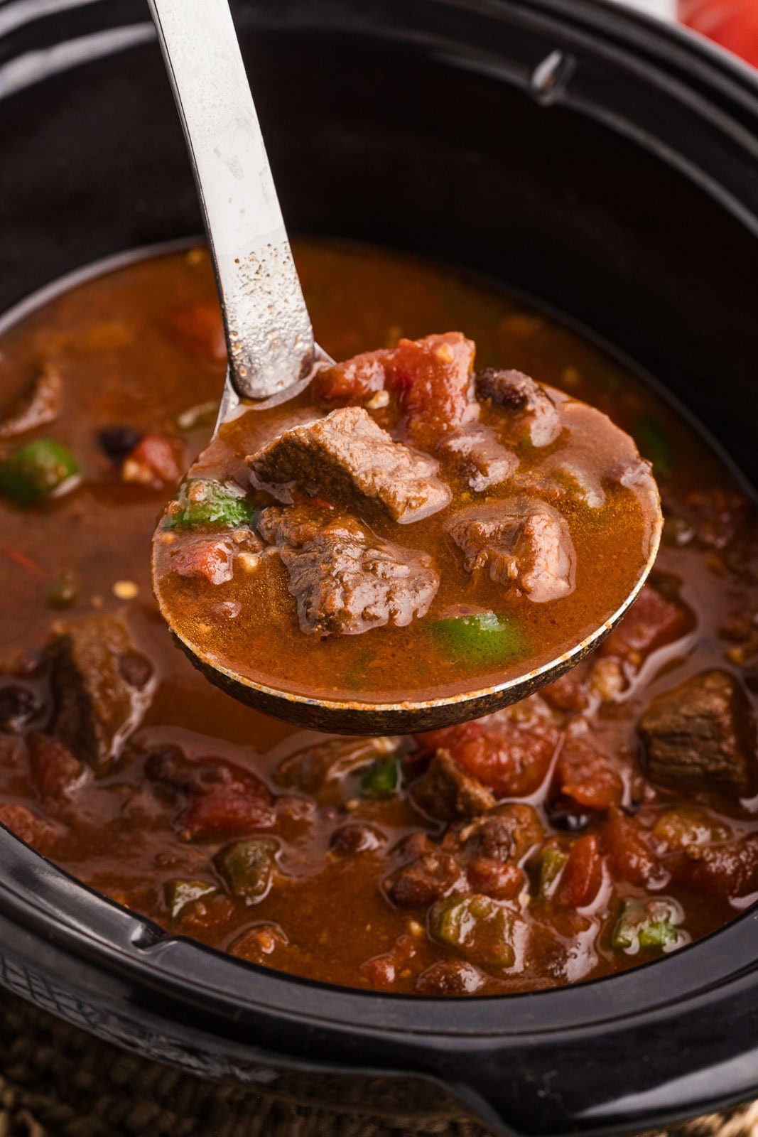 closeup of large ladle containing steak chili over a black slow cooker of chili