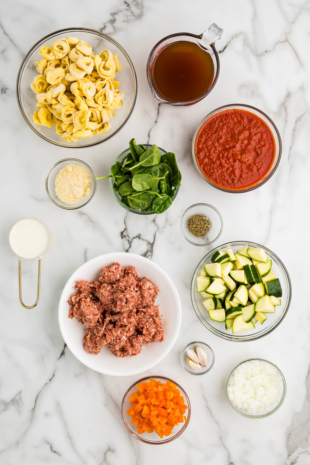 tortellini soup ingredients to include bowls of tortellini, beef broth, marinara sauce, baby spinach, parmesan cheese, heavy cream, ground Italian sausage, seasoning, diced zucchini, diced carrots, cloves of garlic, diced onion