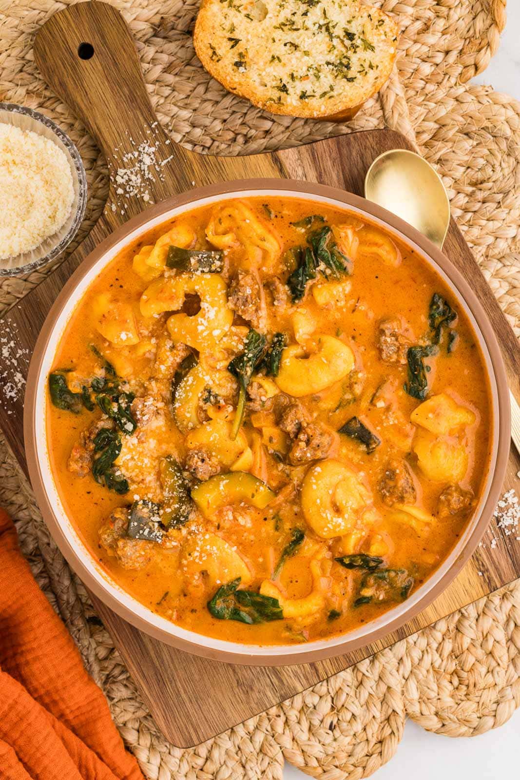 overhead view of bowl of tortellini soup on wooden cutting board and tan placemat, surrounded by gold spoon, bowl of parmesan cheese, and garlic bread