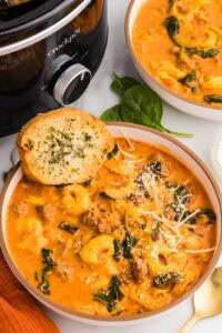 two bowls of tortellini soup, one with garlic bread on side, with black slow cooker in background
