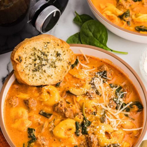 two bowls of tortellini soup, one with garlic bread on side, with black slow cooker in background