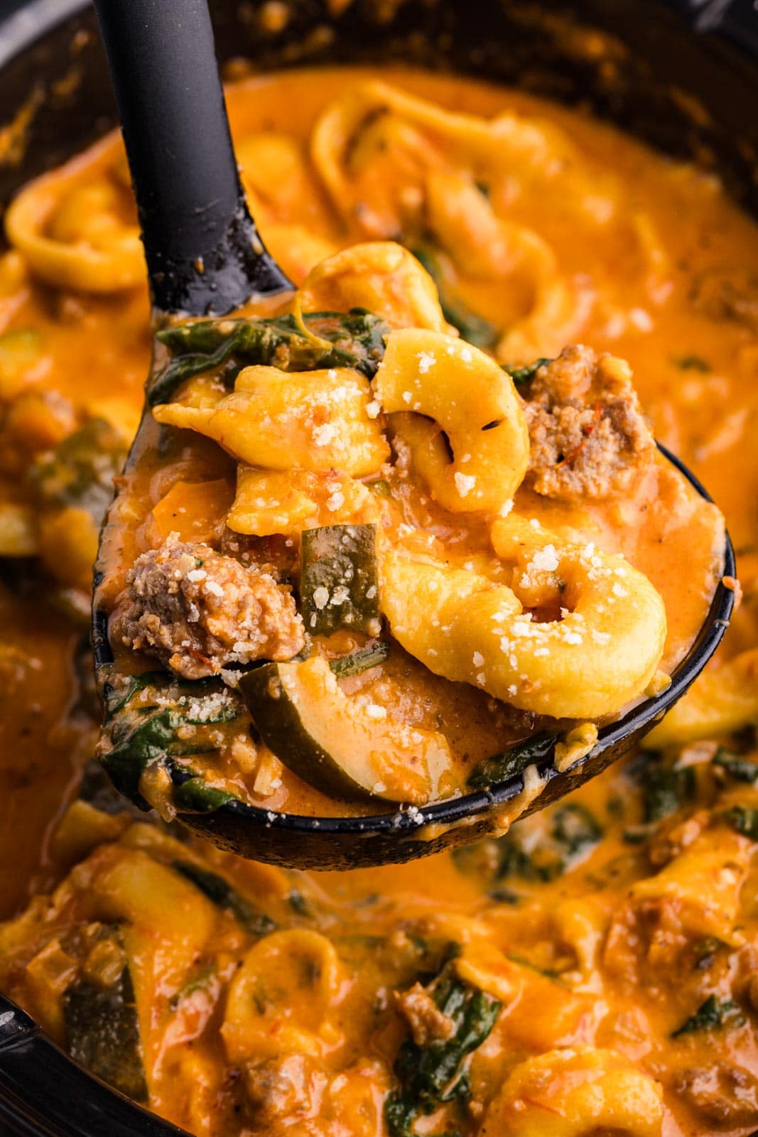 closeup view of black ladle containing tortellini soup with slow cooker of soup in background