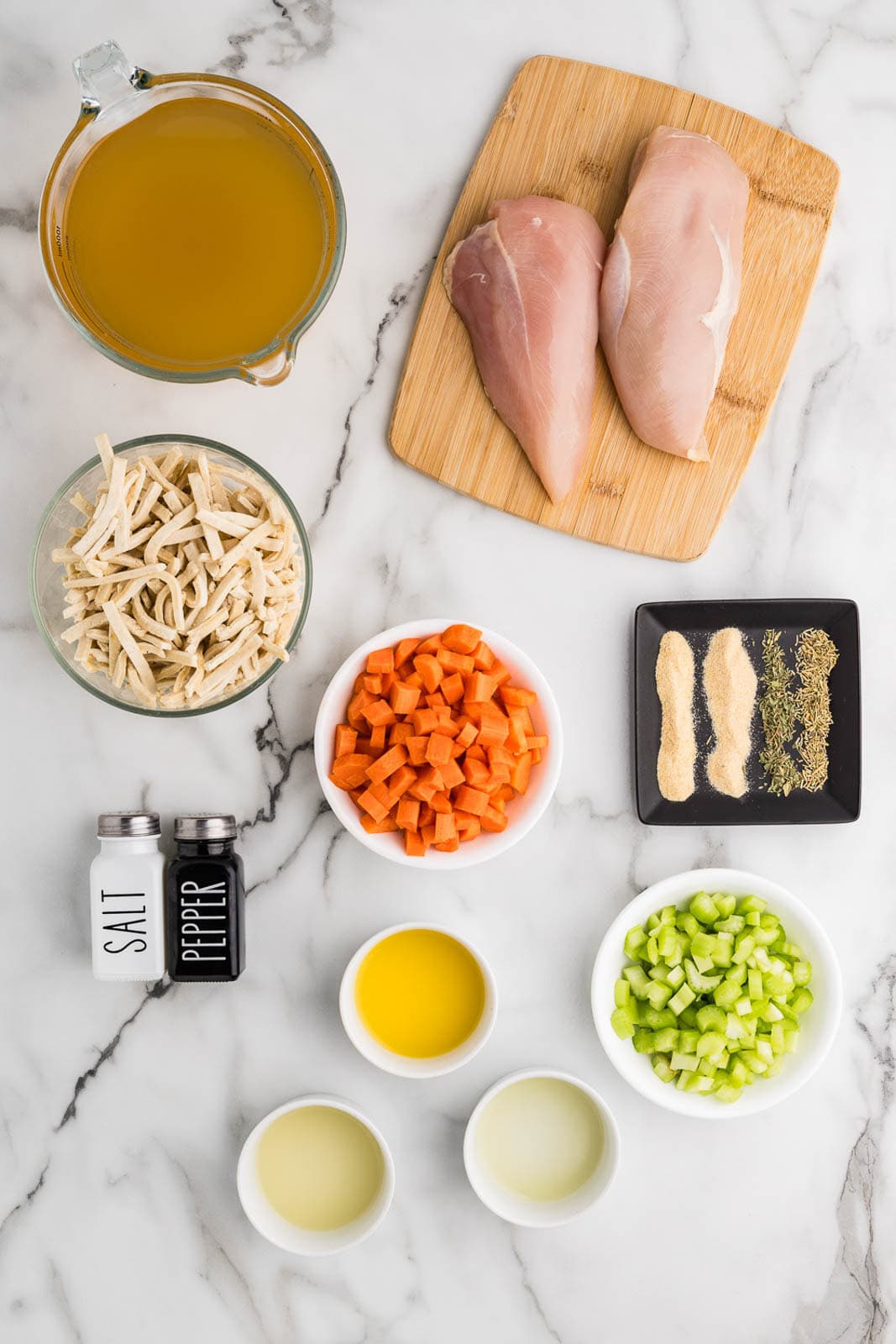 ingredients to include cutting board with two chicken breasts, bowls of seasonings, celery, carrots, frozen noodles, lemon juice, lime juice, orange juice, salt and pepper shakers, and measuring cup of chicken broth