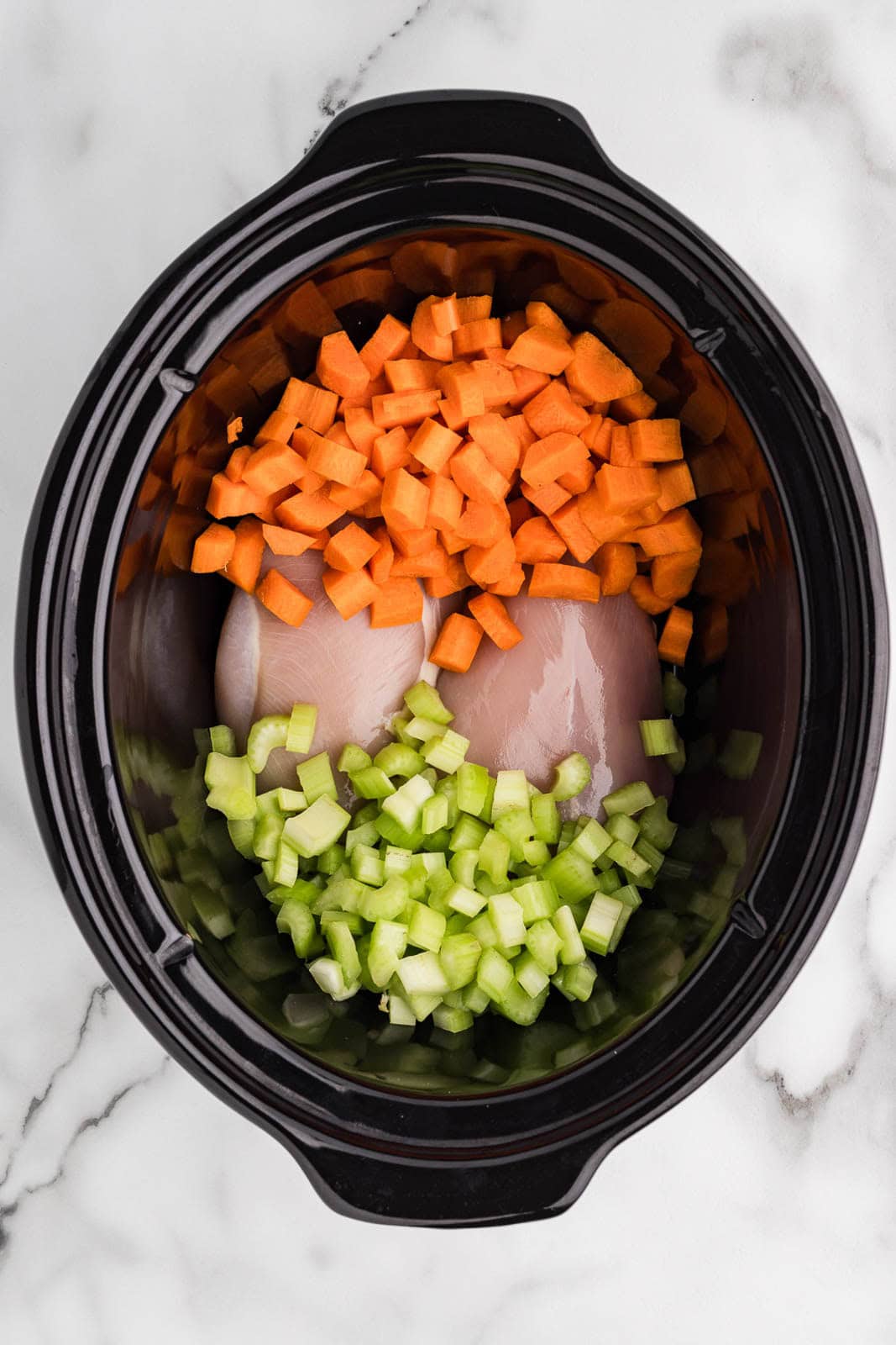 overhead view of black slow cooker containing diced celery and carrots, and two chicken breasts