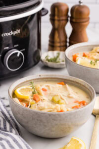 side view of bowl of chicken noodle soup surrounded by wooden salt and pepper shakers, another bowl of soup, gold spoons, and black slow cooker
