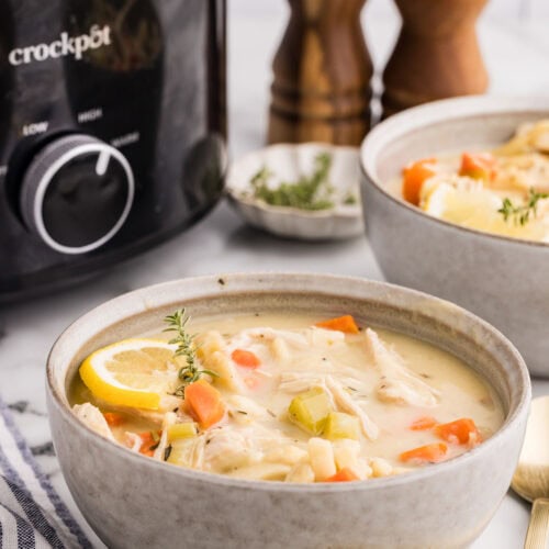 side view of bowl of chicken noodle soup surrounded by wooden salt and pepper shakers, another bowl of soup, gold spoons, and black slow cooker