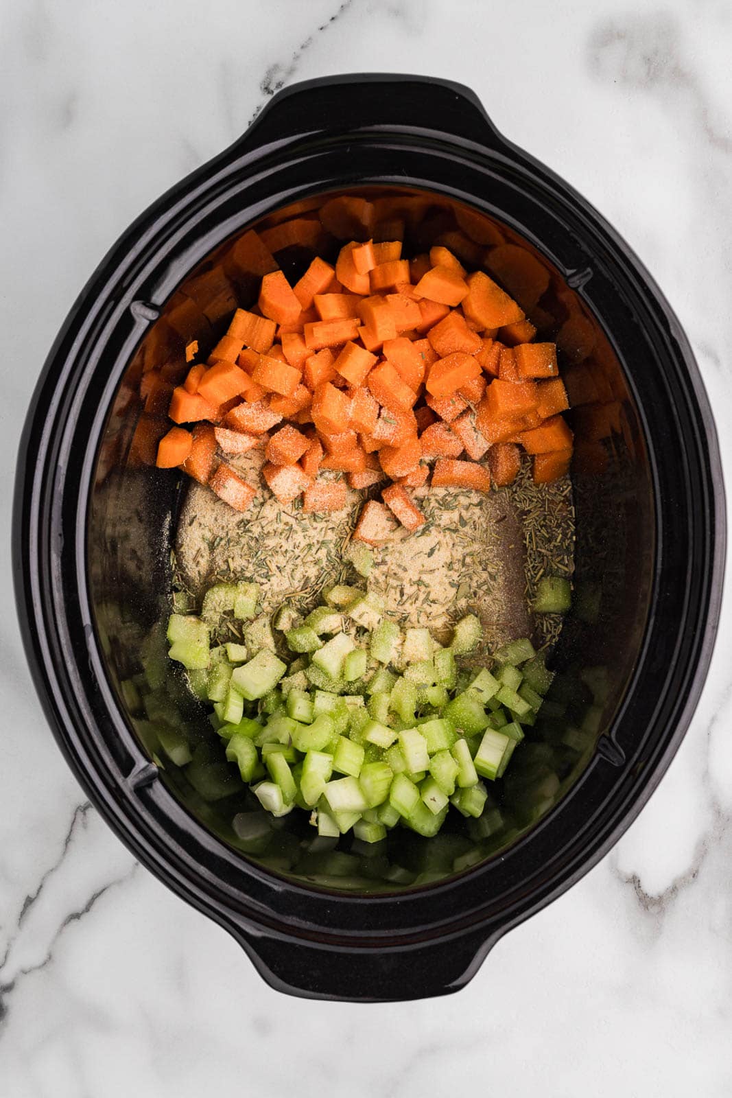 overhead view of black slow cooker containing two chicken breasts, diced celery and carrots, with seasonings
