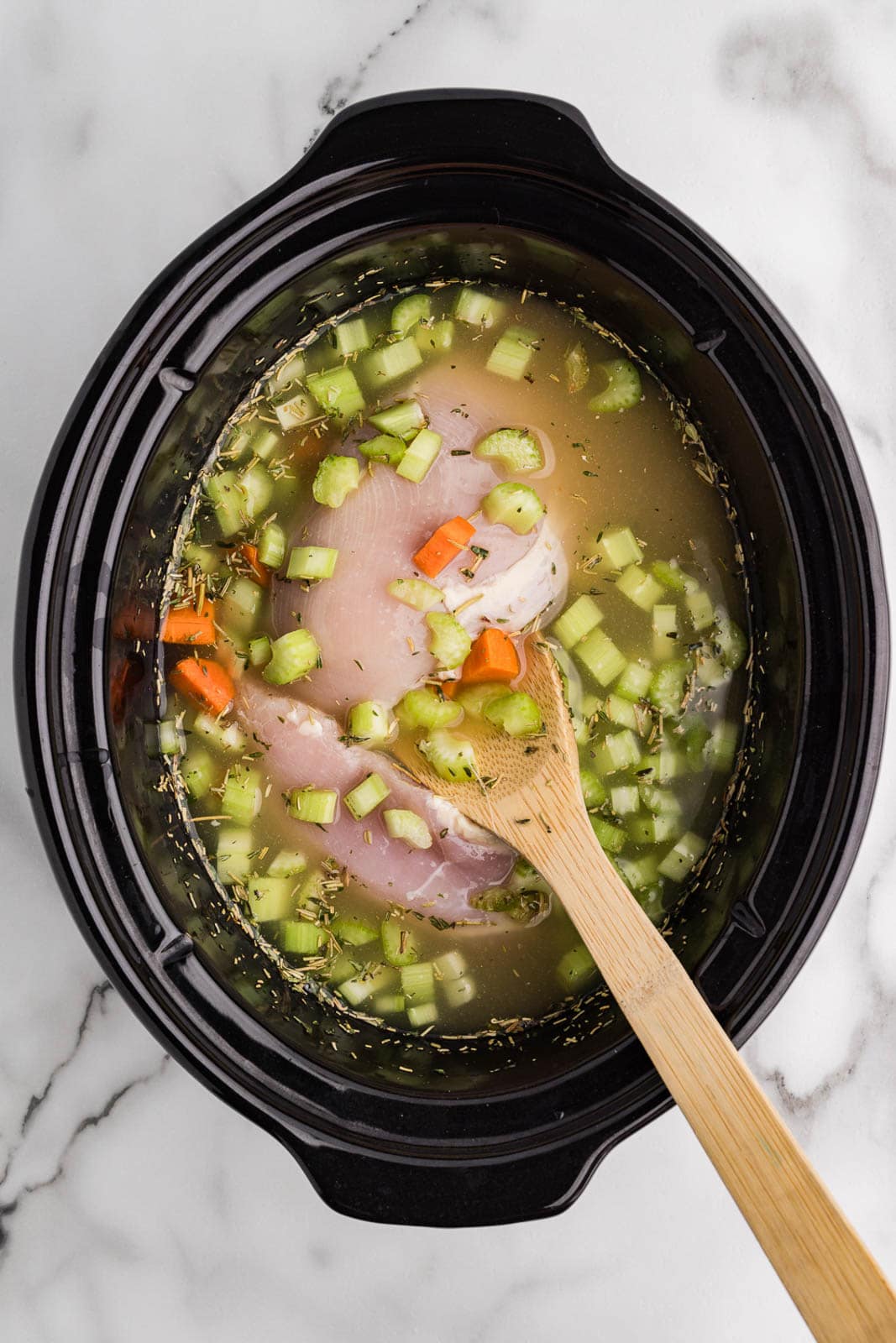 overhead view of black slow cooker containing chicken breasts, carrots, celery and chicken broth, with large wooden spoon