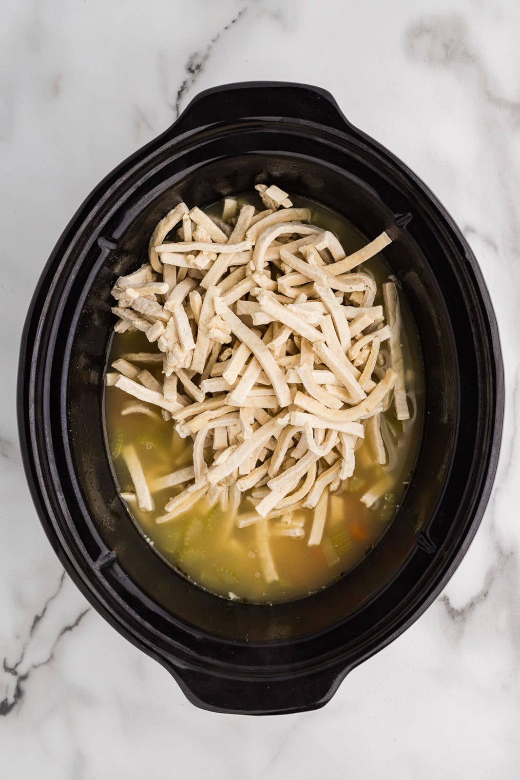overhead view of black slow cooker with chicken soup mixture and frozen noodles on top