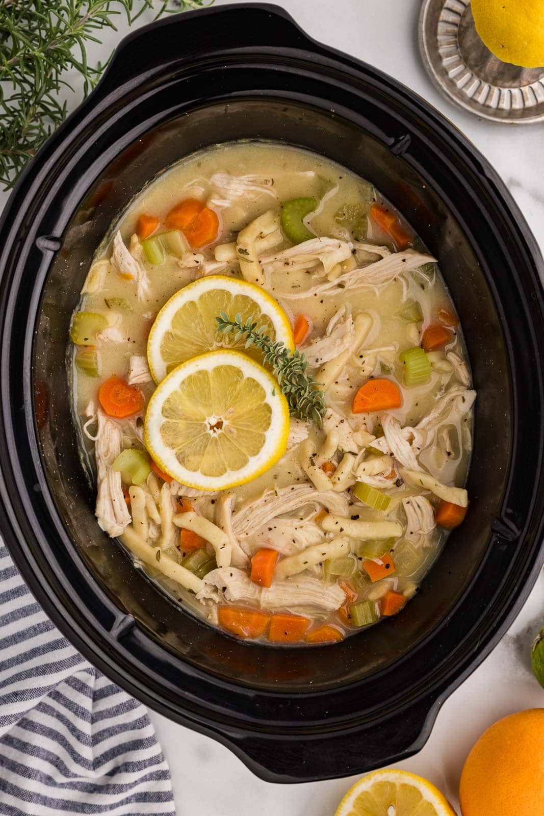 overhead view of black slow cooker of chicken noodle soup with two lemon slices on top, surrounded by oranges and lemons